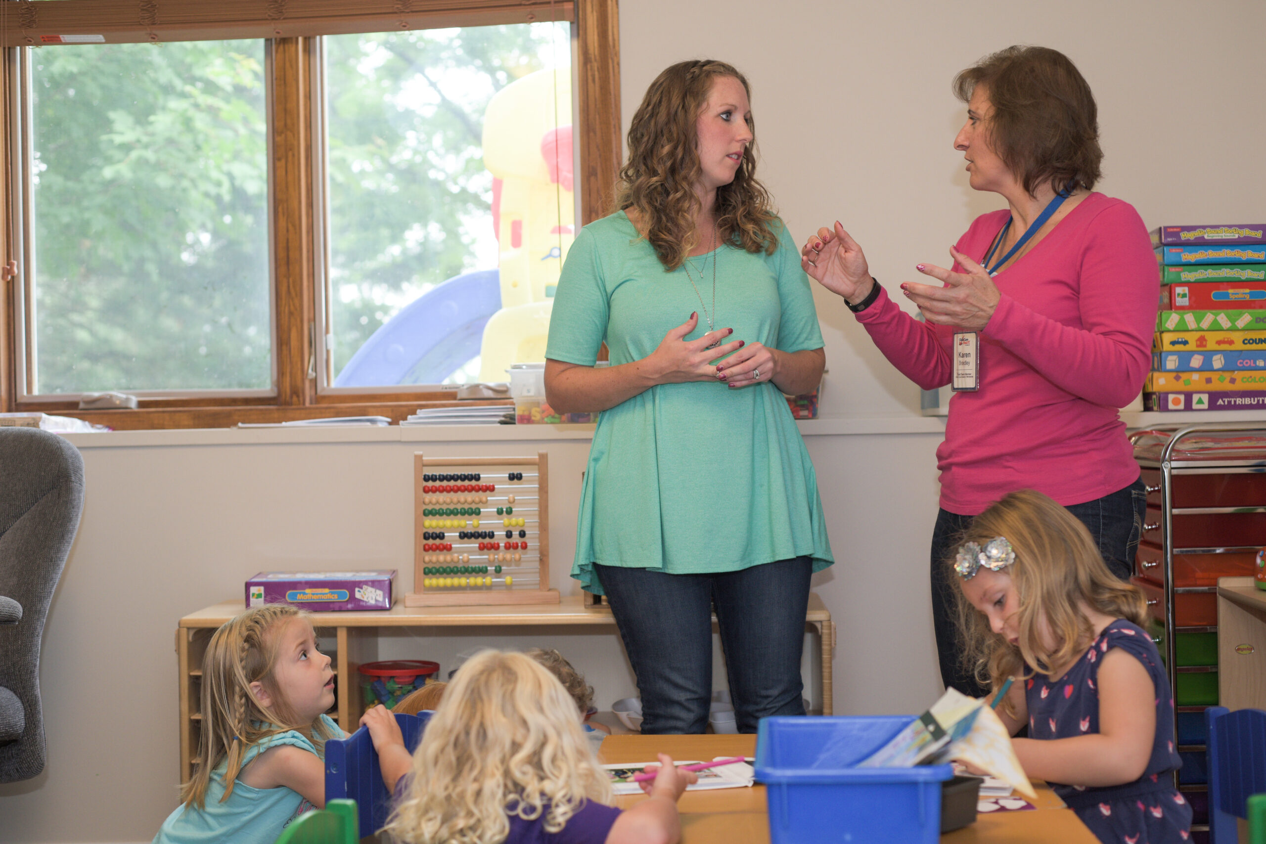 Two adults talk in a classroom while children play